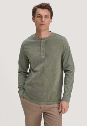 Junger Mann mit langärmligem olivgrünem Henley-Shirt und beigen Hosen, steht mit einer Hand in der Tasche vor einem schlichten Hintergrund.