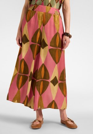 Femme portant une jupe rose avec des formes géométriques marron, orange et beige, associée à des ballerines marron et un bracelet épais.