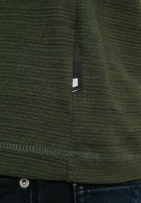 Camisa de tela acanalada color verde oliva con una superficie texturizada, que presenta una etiqueta negra en el dobladillo y un detalle de costura acabada.