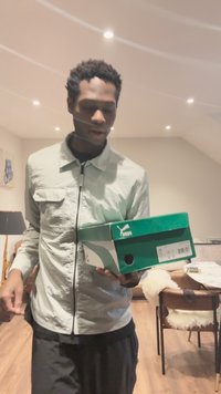 Vyras šviesiu švarku laiko žalią „Puma“ batų dėžutę patalpoje, fone matyti valgomojo stalas ir kėdės.