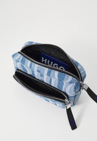 Niebieska kosmetyczka z denimu z abstrakcyjnym jasnoniebieskim wzorem, podwójnymi zamkami błyskawicznymi, czarnym wnętrzem oraz akcentami marki HUGO.