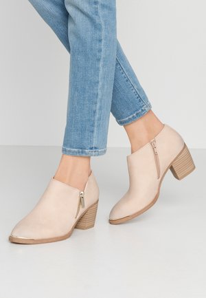 Bottes chevilles beige clair avec fermetures éclair sur le côté et talons empilés portées avec un jean bleu court sur un fond blanc uni.
