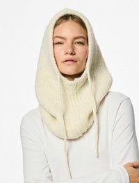 Cappello a maglia color crema con texture a coste, dotato di lacci e dal design oversized e accogliente. Indossato sopra una maglietta bianca a maniche lunghe.