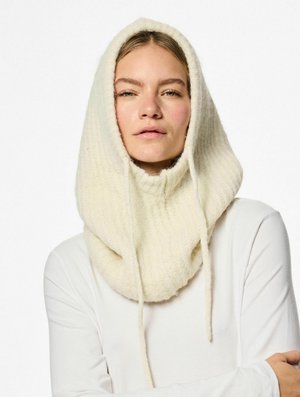 Gorro de punto color crema con textura acanalada, que cuenta con cordones y un diseño oversized y acogedor. Lleva puesto sobre una camiseta de manga larga blanca.