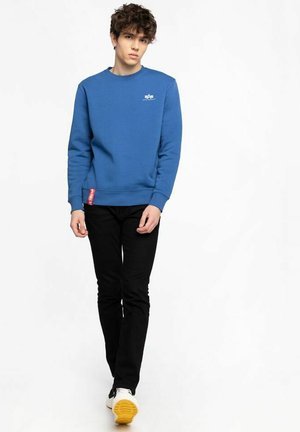 Blauer Sweatshirt mit Rundhalsausschnitt und gerippten Bündchen, versehen mit einem kleinen Logo auf der Brust, kombiniert mit schwarzen Slim-Fit-Hosen und weißen Turnschuhen.