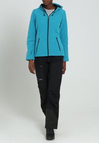 Turkis softshell-jakke med hætte, lynlås foran og to sidelommer, parret med sorte bukser med lommer og kontrastfarver.