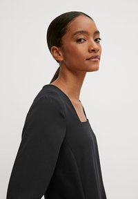 Schwarzes langärmliges Oberteil mit quadratischem Ausschnitt, glatter Textur und figurbetontem Design, das eine minimalistische und moderne Silhouette präsentiert.