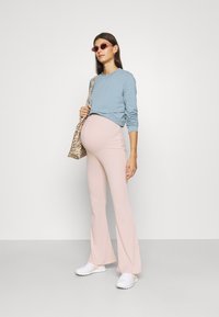 Conjunto de maternidade com um sweatshirt azul claro e calças flare em rosa pálido. Acessórios incluem uma mala de padrão de leopardo e óculos de sol rosa.