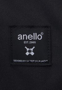 Étiquette en tissu noir avec texte blanc. Indique "anello", "EST. 2005," et "DESIGNÉ PAR CA'POT CO. AU JAPON" avec des accents graphiques.