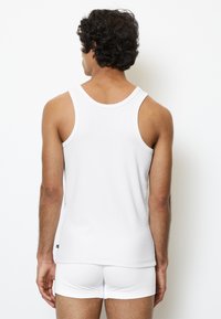 Camiseta sin mangas de algodón blanco con un diseño clásico, que presenta tirantes anchos. Modelo visto desde la parte de atrás, combinado con ropa interior blanca a juego.