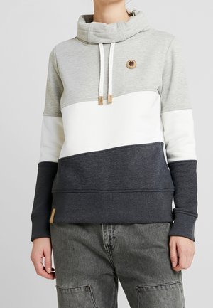 Person iført en farveblok-hættetrøje med grå, hvid og mørkegrå sektioner, med funnelhals og snore, kombineret med grå jeans.