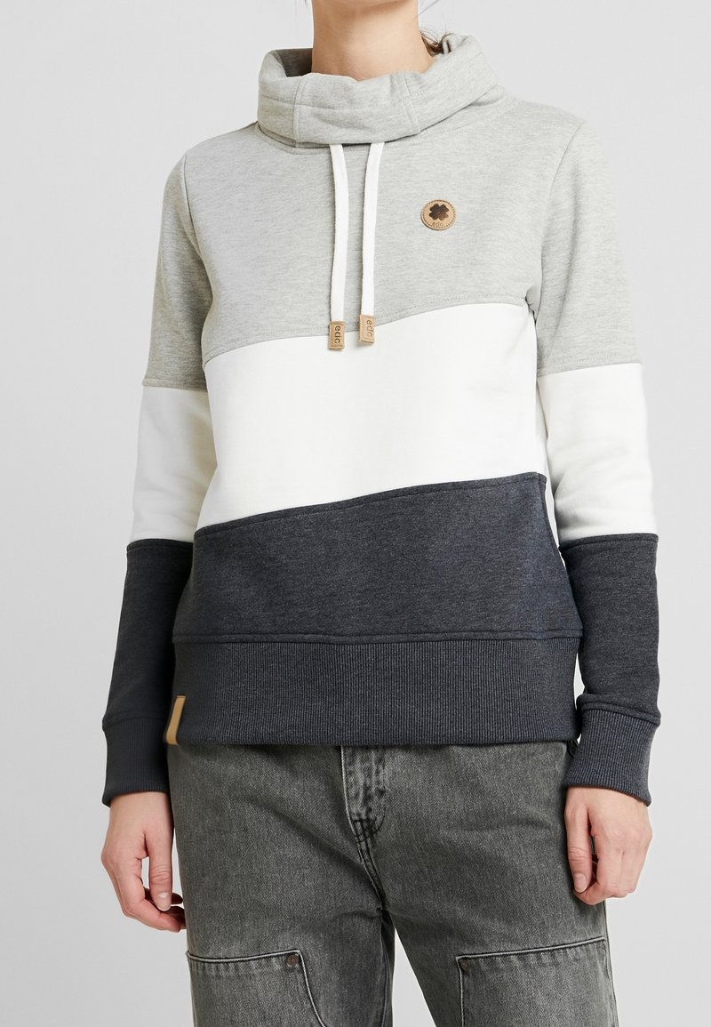 Personne portant un sweat à capuche color block avec des parties grises, blanches et gris foncé, avec un col cheminée et des cordons, associé à un jean gris.