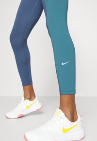 Dwukolorowe legginsy sportowe w ciemnoniebieskim i turkusowym, z białym logo Nike. W zestawieniu z białymi sneakerami z żółtymi akcentami i różowymi detalami.
