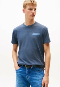 Camiseta de algodón azul marino con cuello redondo, que presenta un logo gráfico en azul claro en el pecho. Combinada con jeans de denim azules y un cinturón negro.
