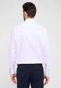 Weißes langärmeliges Hemd mit klassischem Kragen, glatter Textur und Knopfmanschetten, kombiniert mit dunklen Hosen mit zwei Gesäßtaschen.