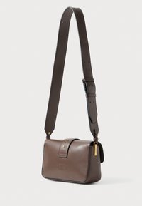 Bolso bandolera de cuero marrón con correa ajustable, tachuelas doradas, logo en relieve y detalle de etiqueta dorada sobre fondo blanco.