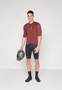 Burgunderfarget sykkeltrøye med korte ermer og frontglidelås. Sorte sykkelshorts. Svart hjelm holdt i hånden. Sorte sykkelsko og sokker.