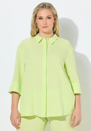 Kvinde iført løs, lysegrøn skjorte med knapper og trekvartlange ærmer samt matchende bukser mod en ensfarvet lys baggrund.