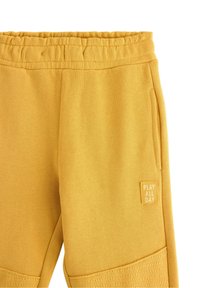 Gule joggingbukser med elastisk talje, sidelommer og en lap med teksten "PLAY ALL DAY". Stoffet har en struktureret overflade.