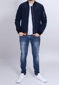 Námořnická bomber bunda s předním zipem, žebrovanými manžetami a oranžovou nášivkou. Kombinována se světlými modrými džíny a bílými teniskami.