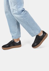 Svarta sneakers med en slät läder- och mockayta, featuring en brun gummisula och mörka snören, bärs tillsammans med ljusblå jeans.