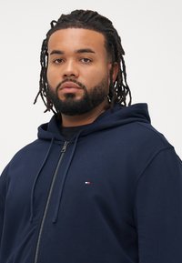Homme avec des cheveux tressés et une barbe, portant un sweat à capuche bleu marine zippé avec un petit logo rouge, blanc et bleu sur la poitrine, faisant face à l'avant.