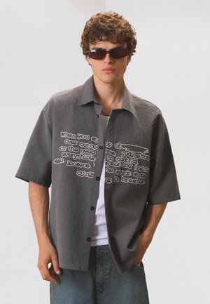 Tânăr purtând ochelari de soare și o cămașă gri cu mâneci scurte, în carouri, cu text alb imprimat, stând cu o mână în buzunar.
