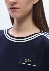 Haut bleu marine côtelé avec col à rayures blanches et petit logo crocodile vert sur la poche. Le tissu semble lisse, design décontracté.