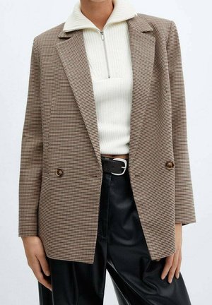 Personne portant un blazer marron à carreaux sur un pull zippé blanc côtelé, un pantalon en cuir noir et une ceinture noire avec une boucle argentée.