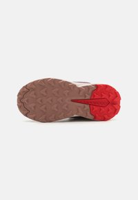 Trail-Laufschuhsohle mit einer braunen Gummi-Basis, die tiefe Traktionsstollen und einen auffälligen roten Absatzakzent aufweist.