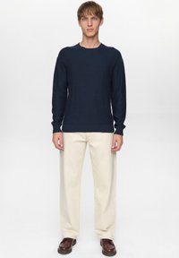 Jonge man met een donkerblauwe trui, crèmekleurige rechte broek en bruine leren schoenen, staand tegen een effen lichte achtergrond.