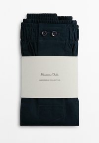 Sous-vêtements bleu marine avec ceinture élastique, dotés de deux boutons et d'un détail de petite poche, enveloppés dans une bande crème texturée avec un logo.