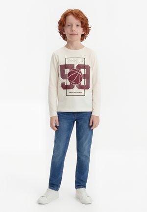 Raudonplaukis vaikas stovi, dėvi kreminės spalvos ilgomis rankovėmis marškinėlį su krepšinio grafika ir skaičiumi 58, mėlynas džinsus ir baltus sportbačius.