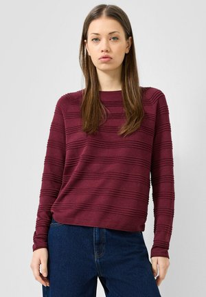 Jonge vrouw met lang bruin haar, die een getextureerde donkerrode top met lange mouwen en donkerblauwe spijkerbroek draagt, staand tegen een effen achtergrond.