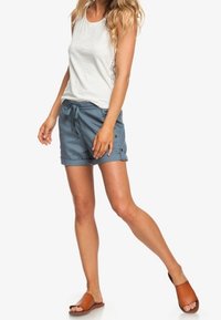 Haut gris clair sans manches avec détail en dentelle, associé à un short bleu avec cordon de serrage et boutons, et sandales marron à enfiler.