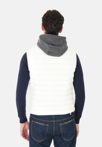 Uomo che indossa un gilet bianco trapuntato senza maniche sopra una camicia a maniche lunghe blu navy e jeans blu scuro, rivolto di spalle su uno sfondo chiaro uniforme.
