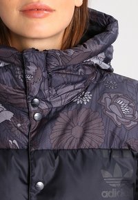 Mörkgrå jacka med blommönster i nyanser av lila och svart. Har en hög krage, metallknappar och en stor logotyp på botten.