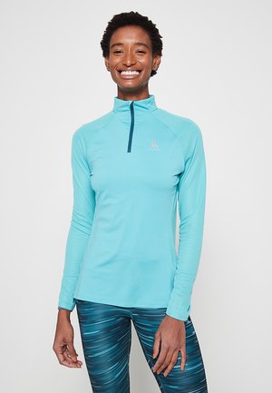 Light blue long-sleeve top with a quarter zip, featuring a smooth texture and fitted design, paired with teal patterned leggings.