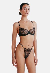 Zwart kanten lingeriesetje met een balconette bh en string, voorzien van dunne verstelbare bandjes en gouden accenten op de hardware.