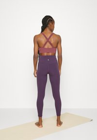 Sutiã desportivo trançado em burgundy com design de costas cruzadas, combinado com leggings roxas de cintura alta com um pequeno emblema nas costas.