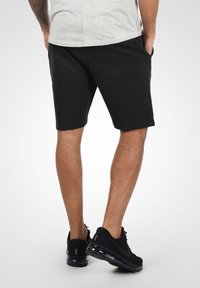 Shorts de sport noirs en tissu doux avec poches latérales. Portés avec des baskets noires dotées d'un amorti à air et de détails en mesh.