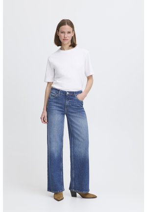 T-shirt blanc à manches courtes associé à un jean en denim bleu à jambes larges. Le jean présente une teinte délavée et une coupe droite. Bottines marron à talons et bouts pointus.