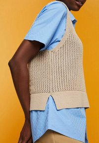 Personne portant une chemise bleu à manches courtes superposée avec un gilet sans manches beige en tricot, sur fond orange.
