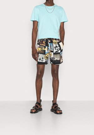 Maglietta di cotone azzurro chiaro, pantaloni corti neri con stampe grafiche miste e accenti gialli, abbinati a sandali neri con cinghie.
