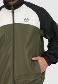 Giacca verde oliva con un design a blocchi di colore bianco e nero, chiusura con zip frontale e tasche laterali. Il materiale presenta una finitura liscia e leggermente testurizzata.
