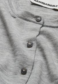 Grijze cardigan van zacht materiaal, met een ronde halslijn, vier zwarte knopen en een label met "ARMEDANGELS" erop.