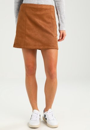 Kobieta w brązowej zamszowej mini spódnicy, szarej bluzce z długim rękawem i białych sneakersach z czarnymi detalami na jednolitym tle.