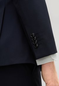 Manica di un abito navy con quattro bottoni neri, tessuto testurizzato e polsino di una camicia bianca che sbuca sotto. Vestibilità sartoriale con linee pulite.