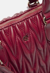 Czerwona skórzana torebka z pikowanym wykończeniem, metalowym zamkiem błyskawicznym i przetłoczonym logo. Posiada gładkie i prążkowane sekcje dla kontrastu.
