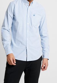 Chemise à boutons bleu clair à rayures verticales blanches. Comprend une poche poitrine et un petit logo. Fabriquée en coton avec une texture lisse.